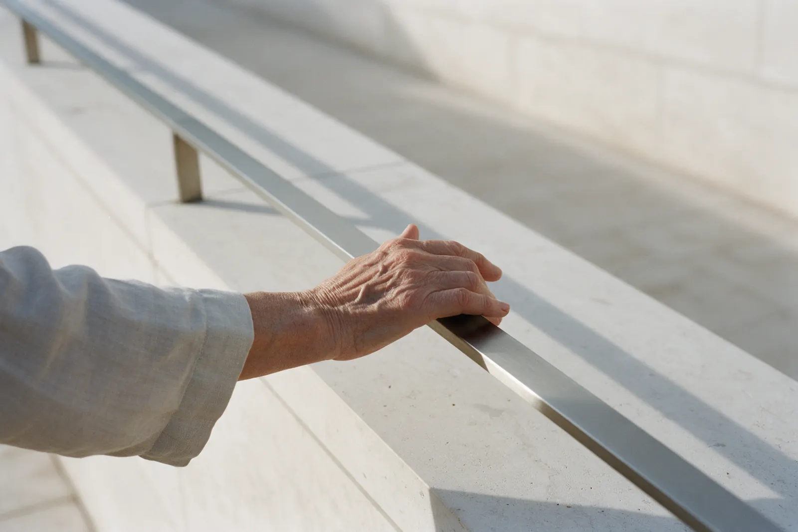 An older person’s hand on a steel handrail running along a limestone ramp, in morning light.